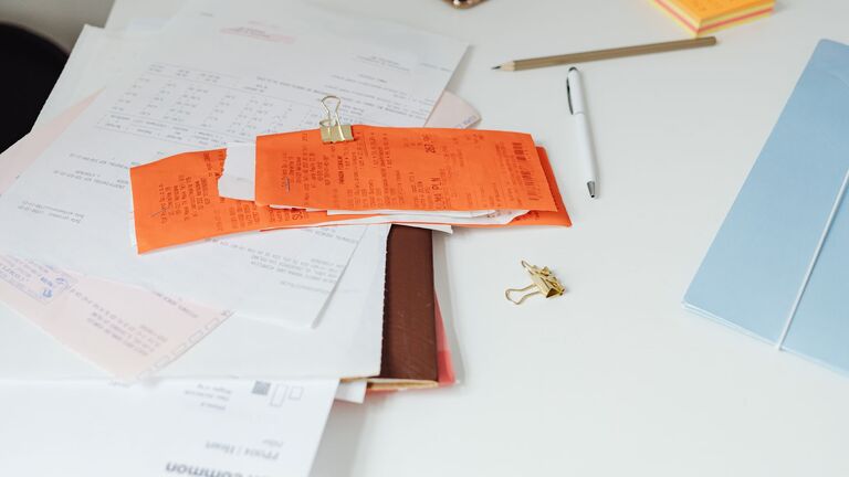 Facturas y tiques sujetos con una pinza sobre un escritorio blanco junto a carpetas, un bolígrafo y notas adhesivas