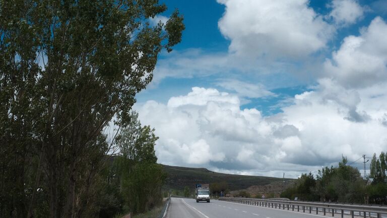 Camión circulando por una carretera de dos carriles entre árboles y lomas bajo un cielo con nubes, en un viaje de larga distancia