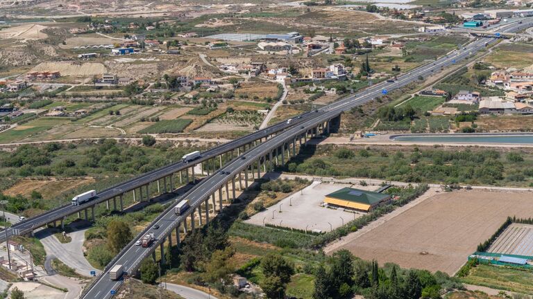 Vista aérea de una autovía en viaducto con camiones que cruza huertas, pueblos y lomas secas de tierra parda