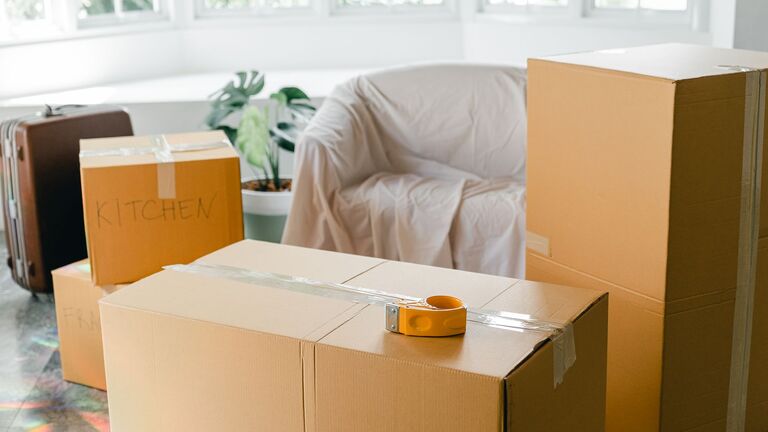 Cajas de cartón cerradas y rotuladas, una maleta y un sillón cubierto con una sábana en un salón luminoso listo para la mudanza